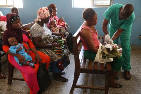 A doctor in green administers care to a baby in a room where many women and babies are sitting.